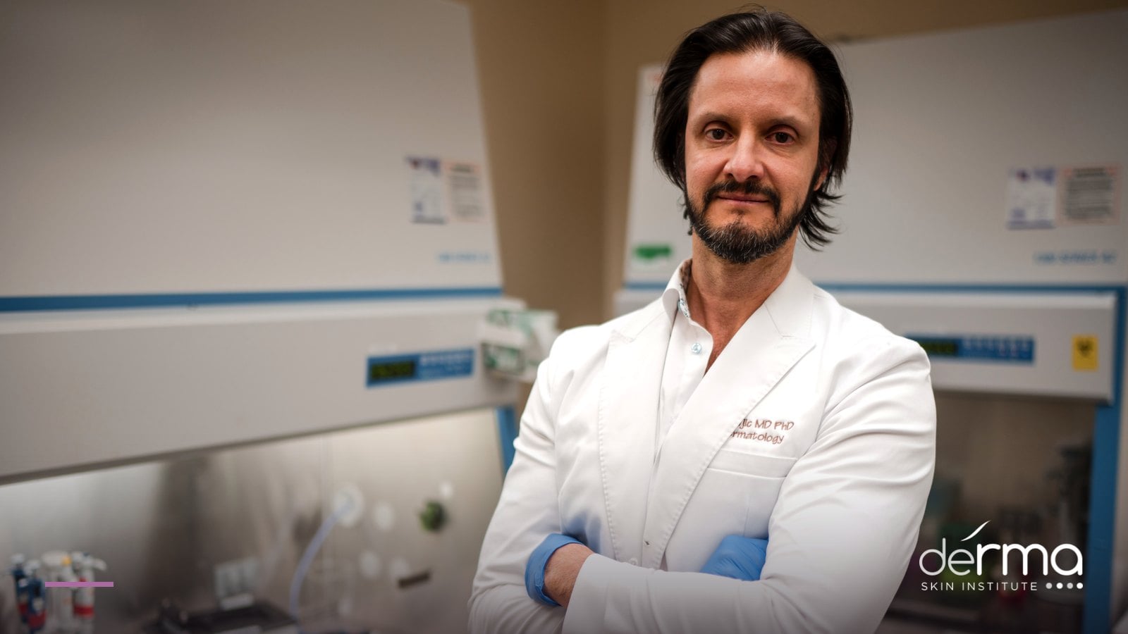 Dr. Dusan Sajic in a white coat embroidered MD PhD Dermatology, standing in a research lab
